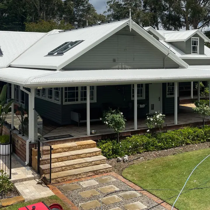 Grey weatherboard home with a pale metal roof and a wide verandah after cleaning, with a hose on the lawn.